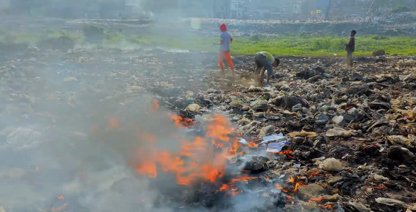 Image: Children walk across a waste dump, collecting salvageable pieces, while a fire sends polluted smoke in the air. Photo courtesy of VOICE.