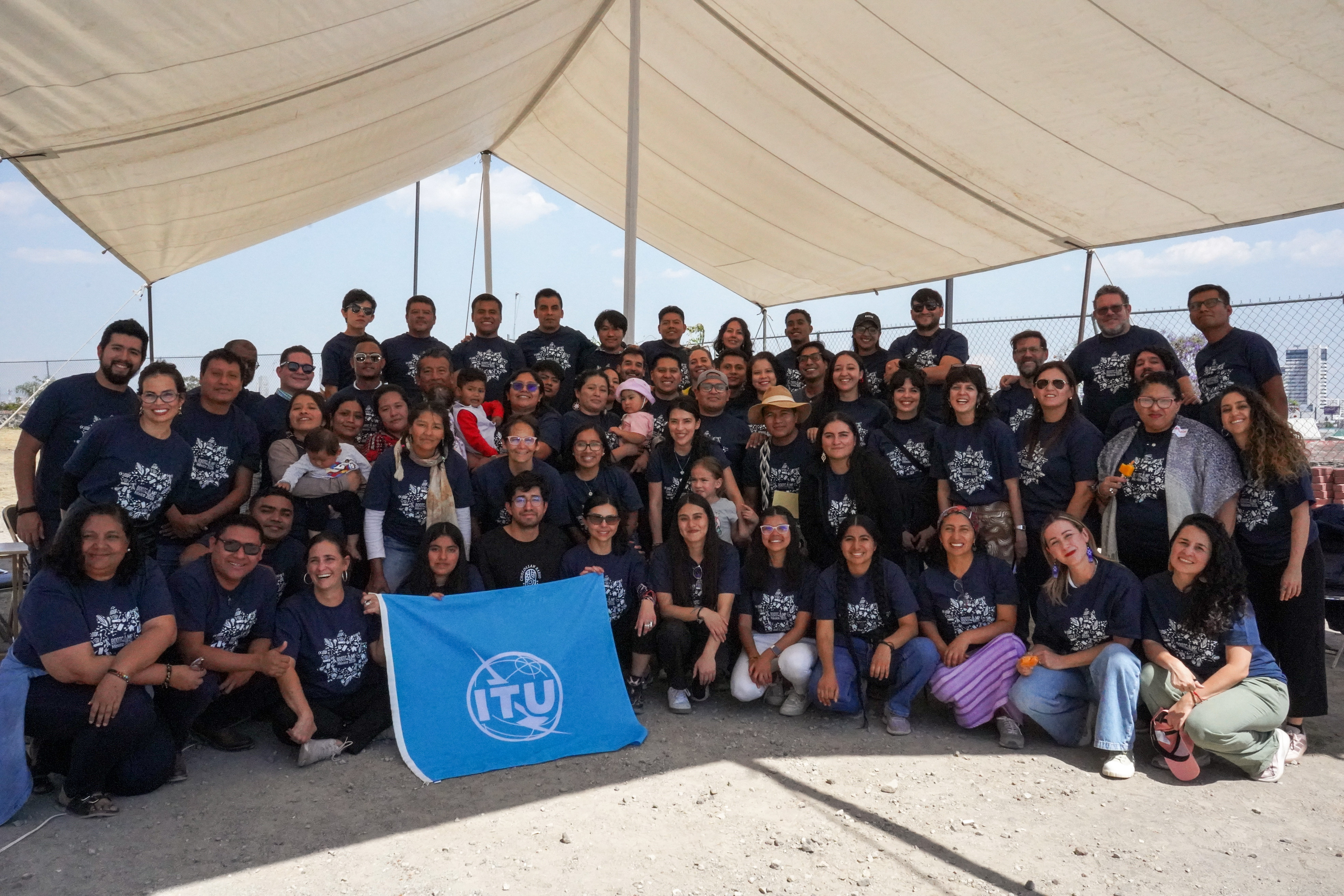 grupo numeroso de personas posando para la foto, todos con la misma remera, sosteniendo una bandera de la ITU