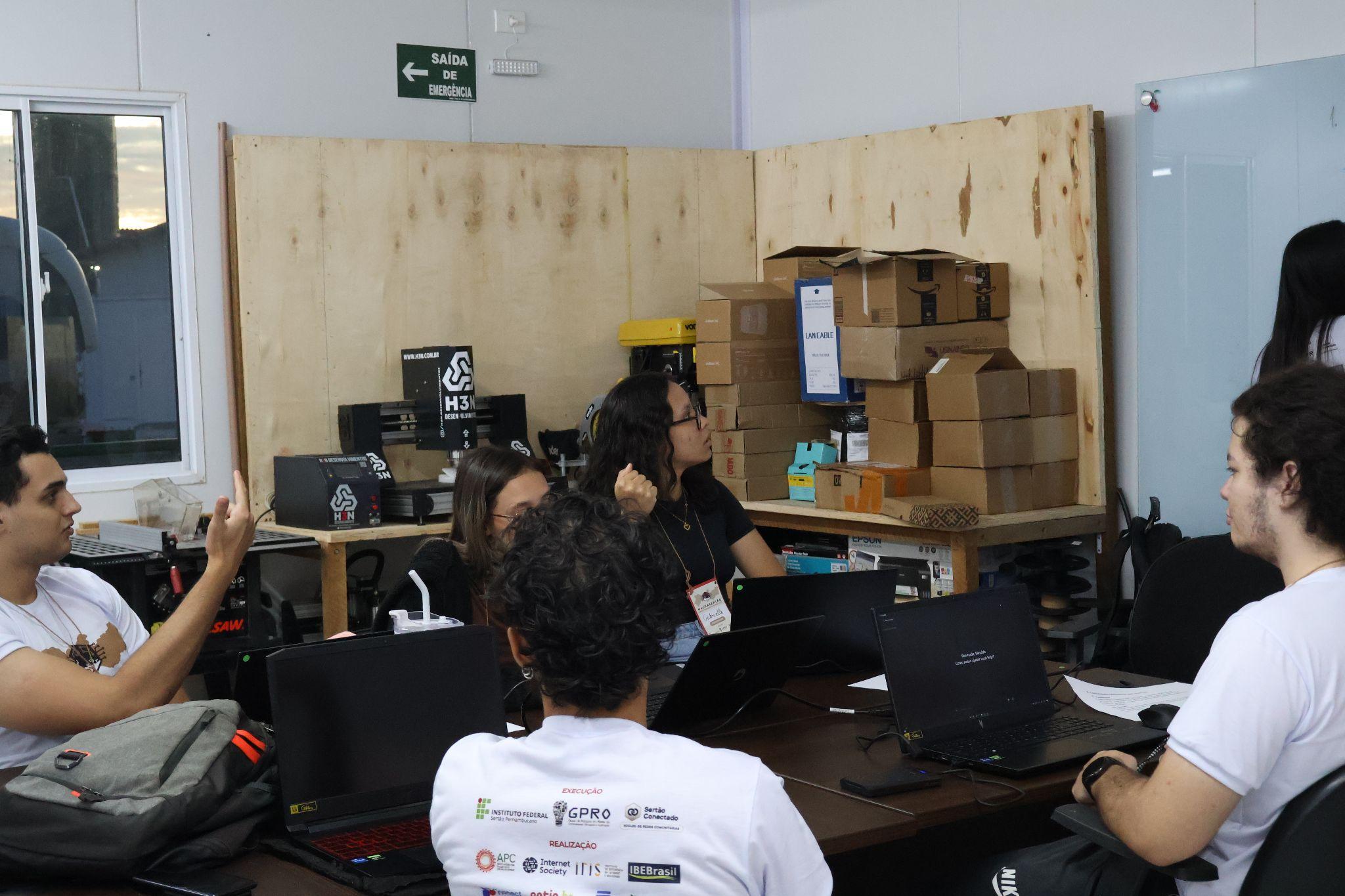 A group of young people are gathered around a table in a workshop or lab setting, working with laptops. Several cardboard boxes are stacked on shelves in the background, and a wooden partition separates the space. One person on the left gestures while speaking, and others listen attentively. Everyone is casually dressed, some wearing white t-shirts with logos on the back. A green “Saída de Emergência” (Emergency Exit) sign is visible on the wall.