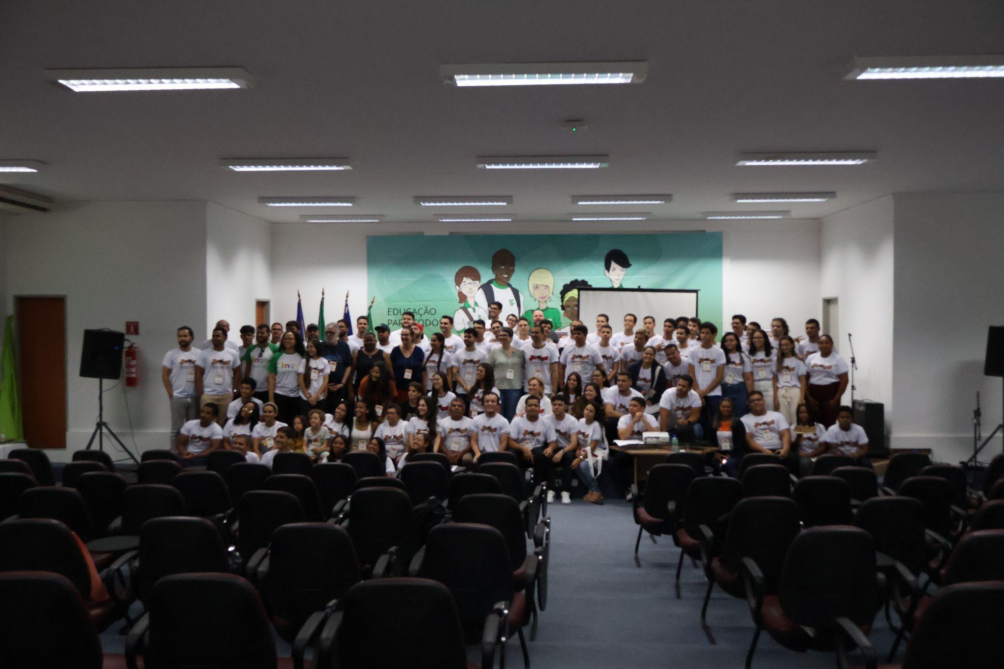 A large group of people poses for a group photo in an auditorium. Most are wearing white t-shirts with logos, and some wear name tags. Behind them is a green banner with cartoon-style illustrations of diverse students and the text "Educação para todos" (Education for all). Flags stand to the left of the stage, and empty rows of chairs fill the foreground.