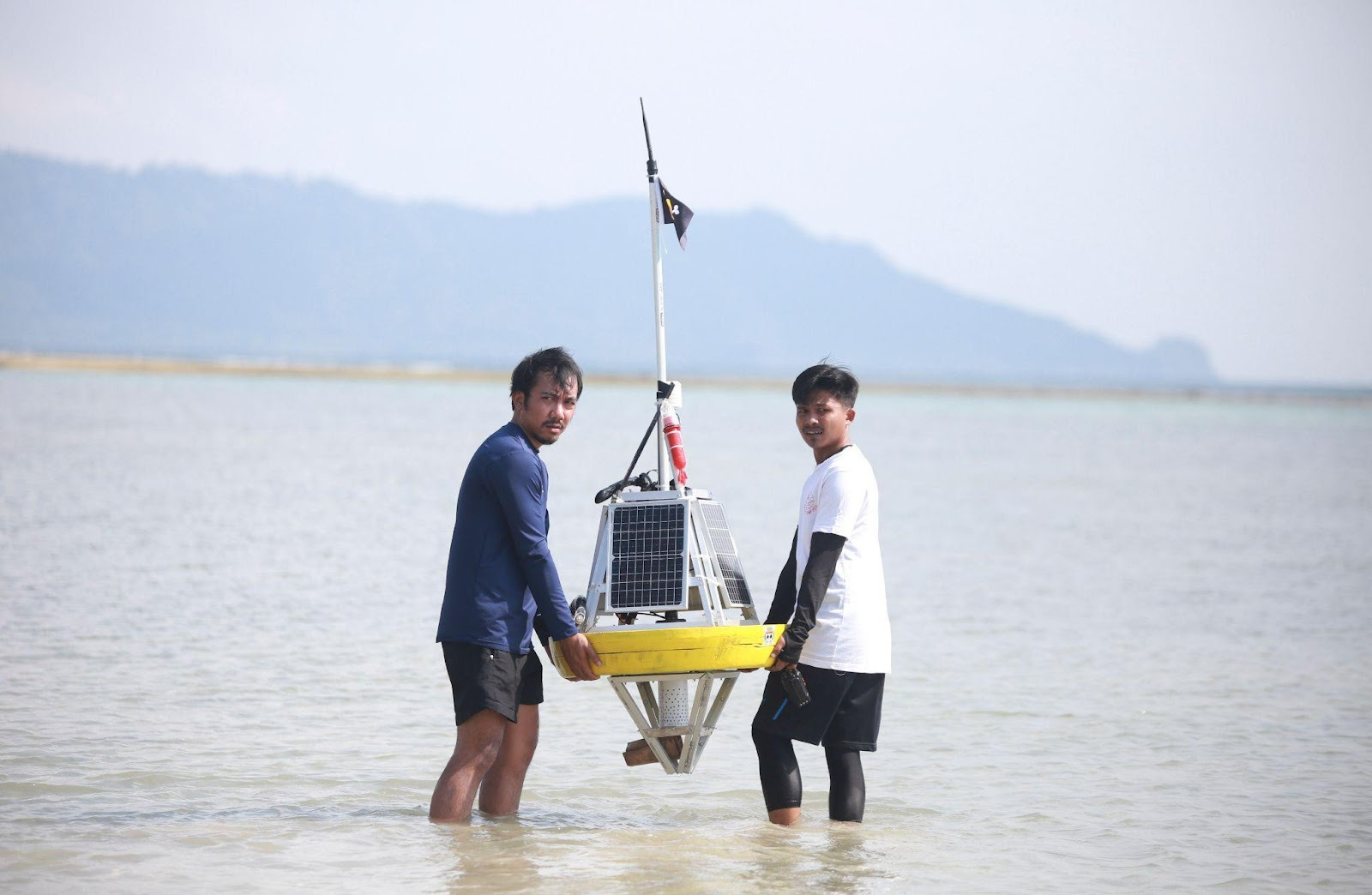 Two men in the water holding a climate sensor device. A moutain is seeing in the other coast of the water.