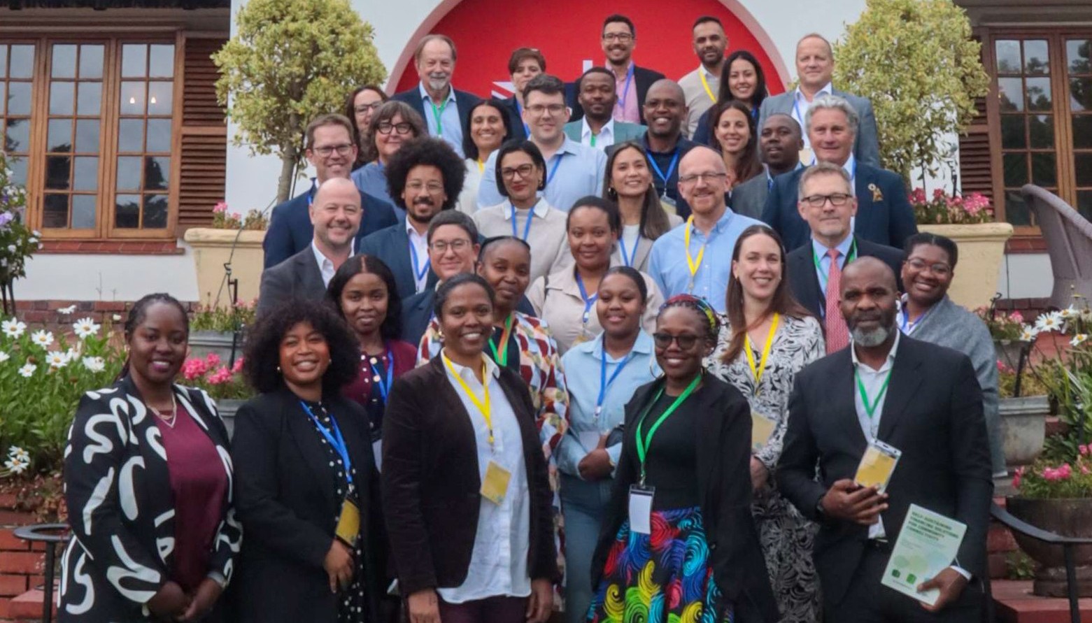 A large group of people poses together on outdoor steps in front of a building with a red and white arch and wooden windows. The group includes men and women of diverse backgrounds, smiling and wearing conference badges on lanyards.
