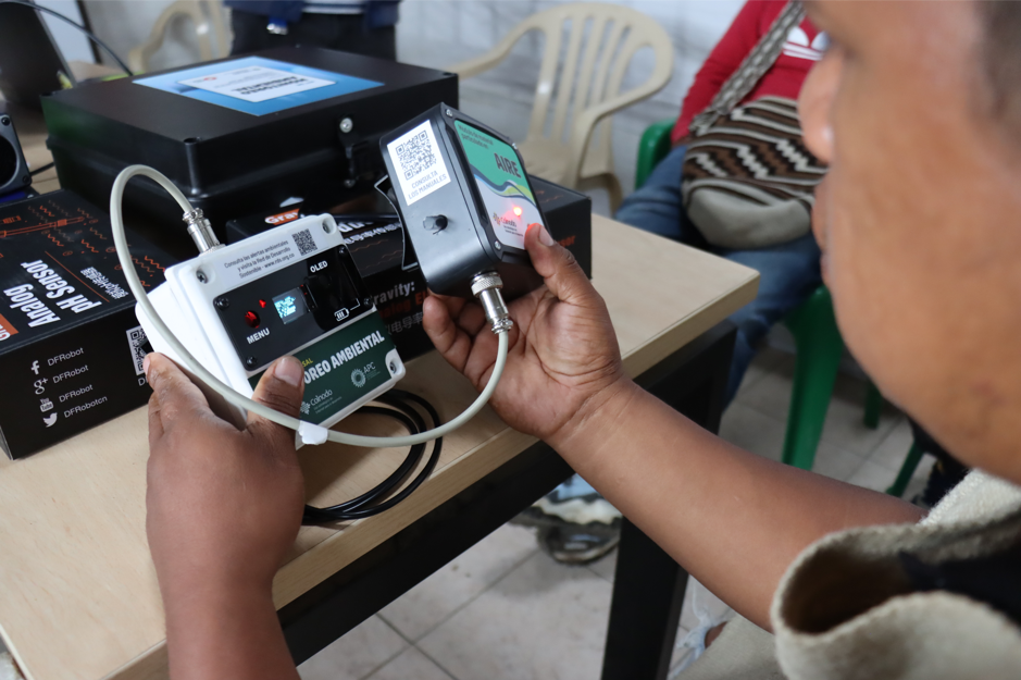 A person holds and inspects air quality monitoring devices connected by cables on a table, with other people and chairs visible in the background.