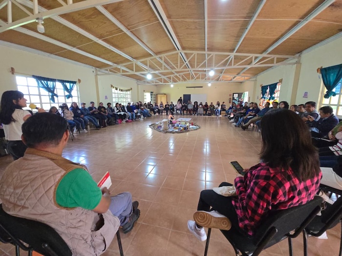 In a large room, dozens of people gather in a circle during the meeting. In the centre, objects brought by the participants as offerings for this collective moment can be seen.