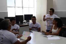 A group of people sitting around a table in a classroom setting, discussing and working together. Several participants wear white T-shirts with the word “Hackasertão.” One person stands and gestures while speaking, while others sit with laptops, papers, and notebooks.