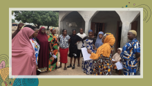 A group of women stand together outside near a building, forming a circle as one woman speaks and holds papers, suggesting a community meeting or training session.