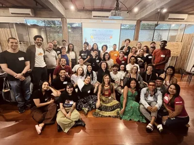 A diversified group of about 50 people smile to a camera in a room. Behind them, a cloth displays a text about connectivity in the communities. In a lively atmosphere.